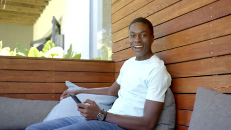 young african american male sitting, holding smartphone, smiling, at home