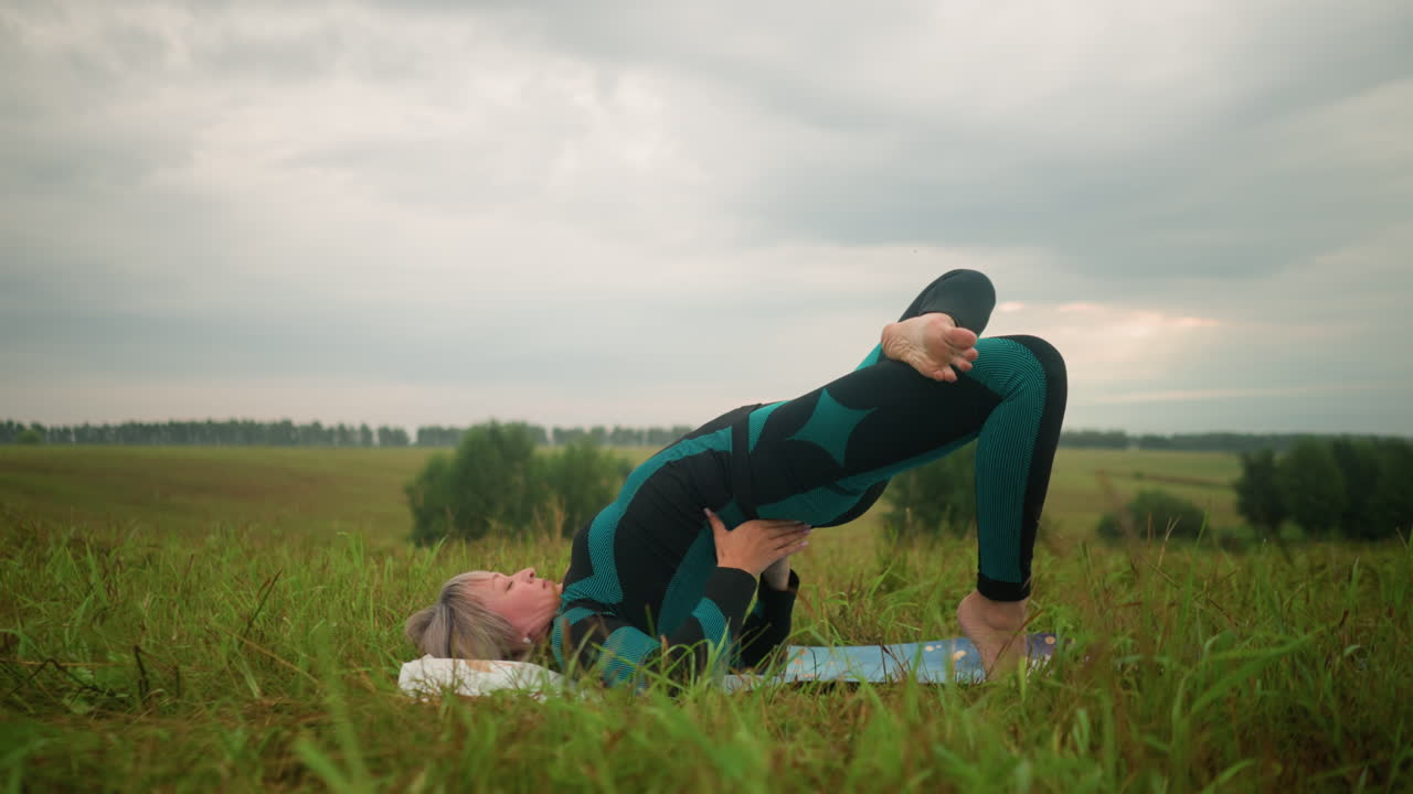 mujer de mediana edad en traje negro verde acostada en una alfombra de yoga practicando la postura del puente con las manos apoyando su cintura, rodeada por un vasto campo de hierba bajo un cielo nublado, con árboles en la distancia