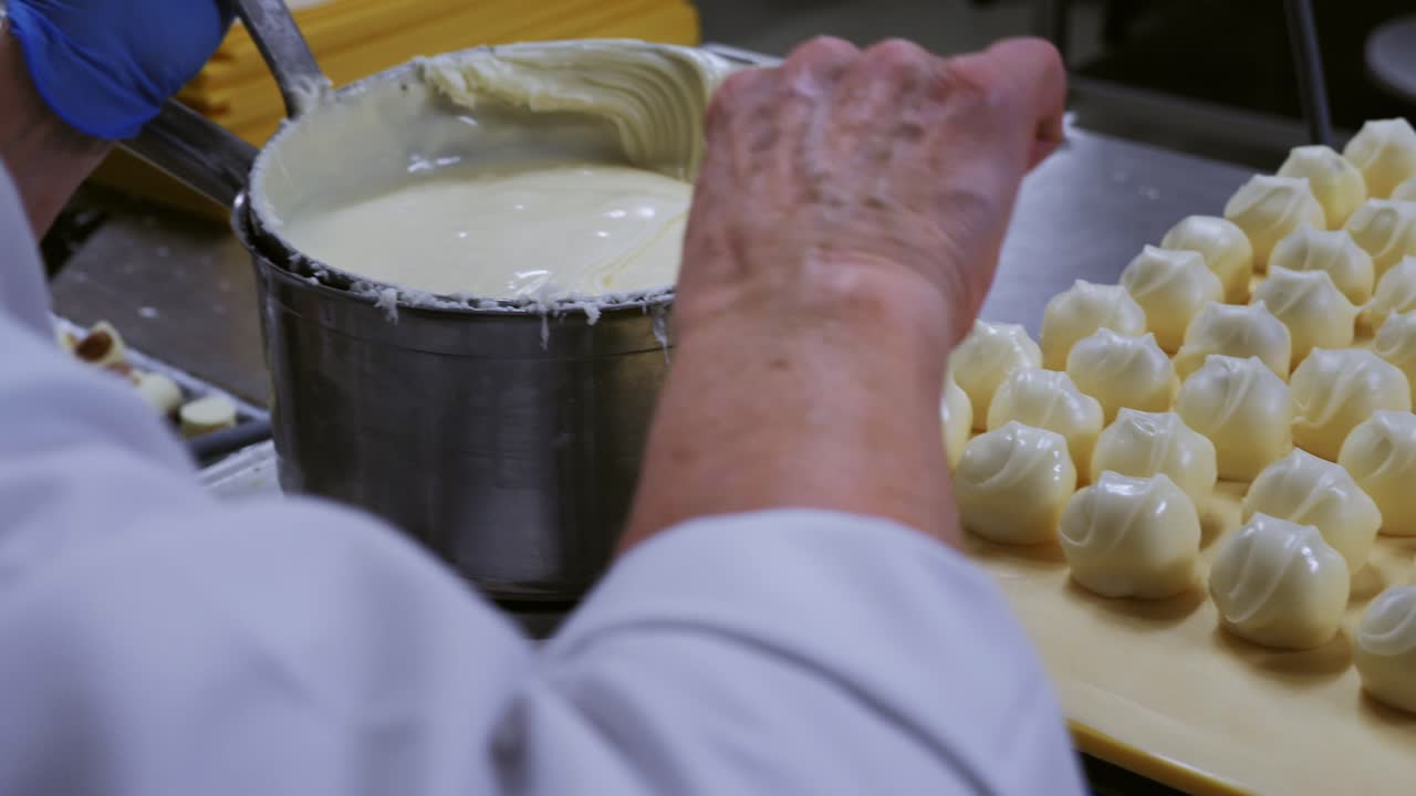 Close-up of worker in white coat and blue gloves scooping white chocolate cream beside finished pralines