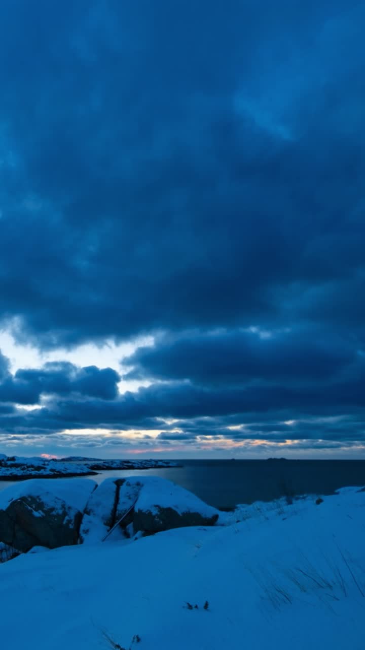 Le nuvole si muovono rapidamente sopra le baite sulle piccole isole innevate vicino al fiordo nella blue hour del primo mattino. 9:16 Verticale video 4K in time-lapse. Isole Lofoten, Norvegia settentrionale