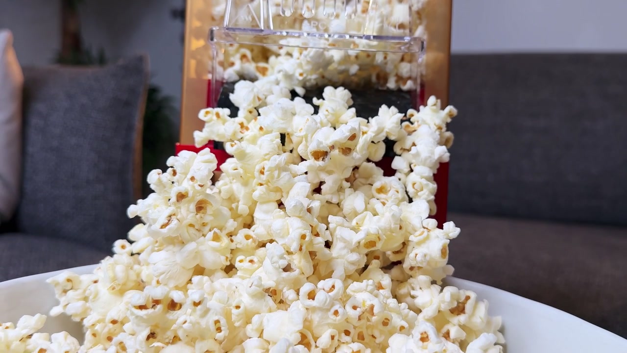 Primo piano di popcorn freschi che cadono da una macchina per popcorn domestica in una ciotola bianca. Ambientazione interna di un soggiorno con illuminazione serale calda per un concetto di spuntino da serata film.