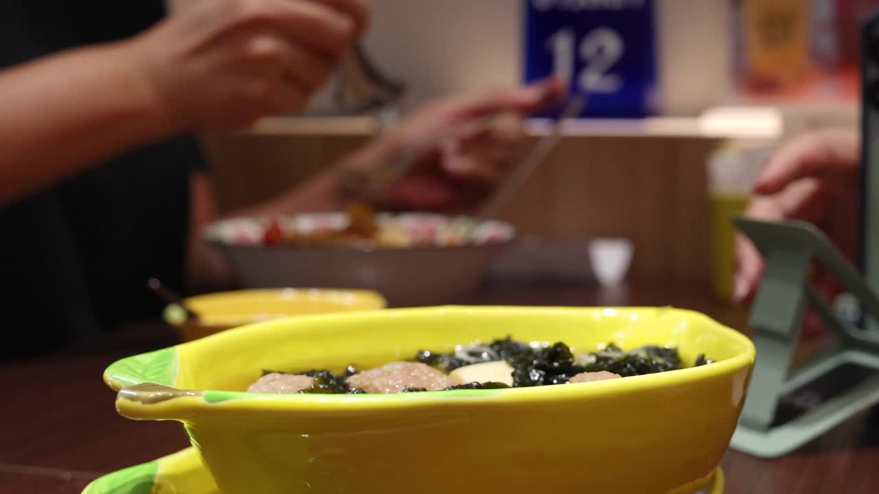 A person uses a spoon to eat pork ball and seaweed soup from a yellow bowl in a casual Thai restaurant with warm indoor lighting and shallow depth of field