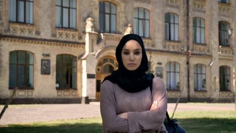 young self-confident muslim girl is standing with arms crossed in daytime in summer, watching at camera, building on background, religious concept