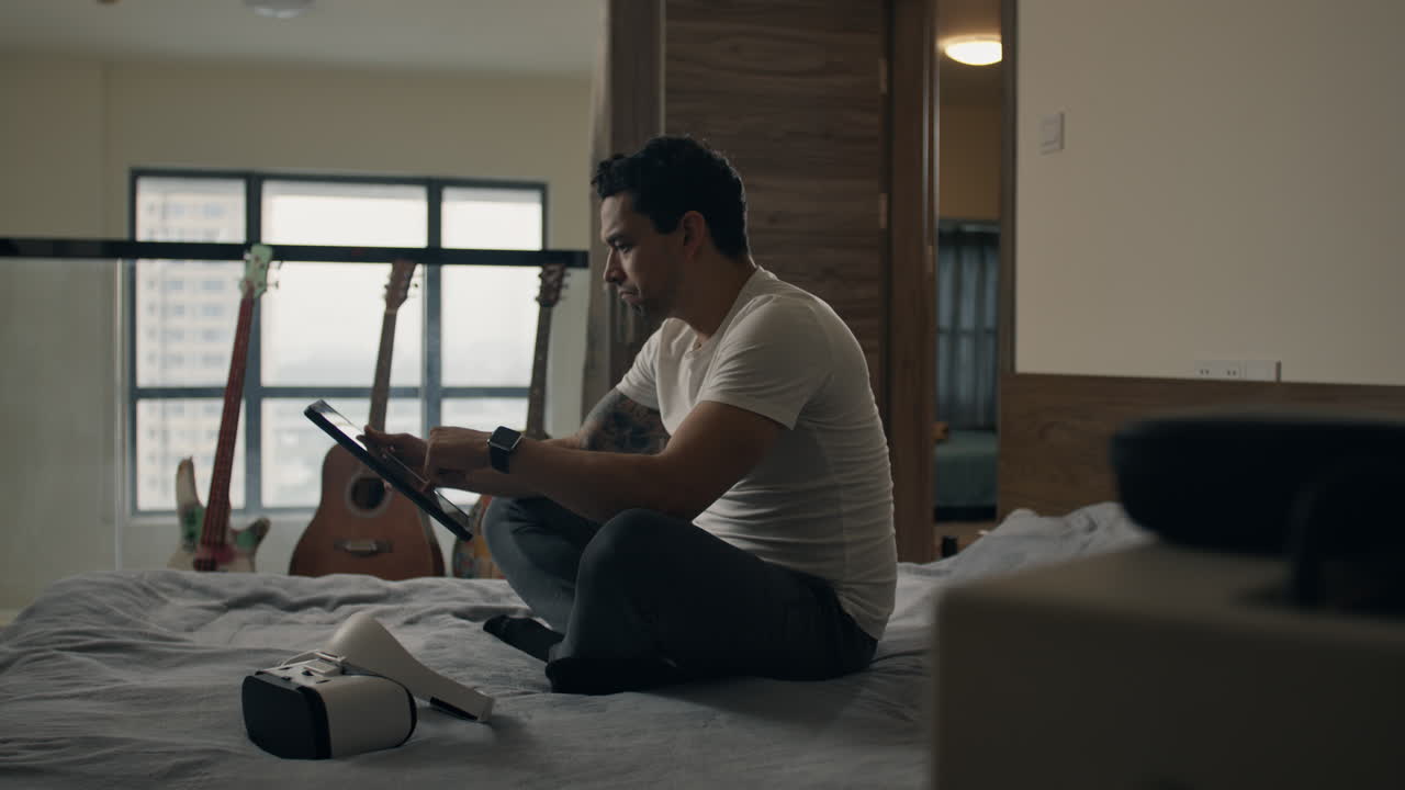 Relaxed man using tablet and VR headset at home, sitting on bed with guitars nearby