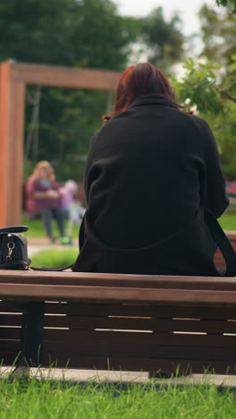 back view of lady sitting on park bench, dressed in black coat and boots, surrounded by lush green trees, relaxed outdoor atmosphere in urban park, blurred view of two people on swing chairs