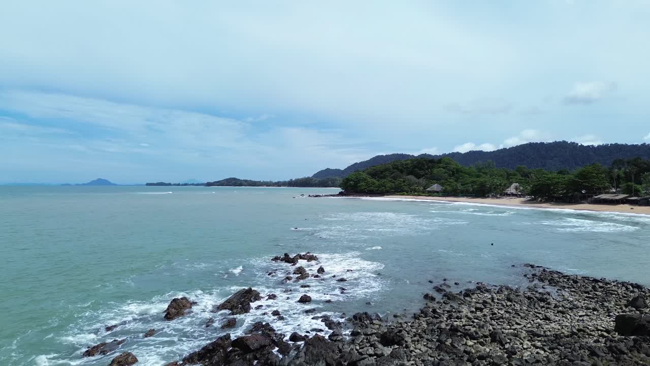 ฟุตเทจดรোনทางอากาศแสดงให้เห็นชายหาดเขตร้อนที่เป็นหินบนเกาะลันตา ประเทศไทย โดยมีต้นเตยเป็นฉากหน้าและทะเลสีเทอร์ควอยซ์ที่เงียบสงบเป็นพื้นหลังภายใต้ท้องฟ้าที่มีเมฆมาก