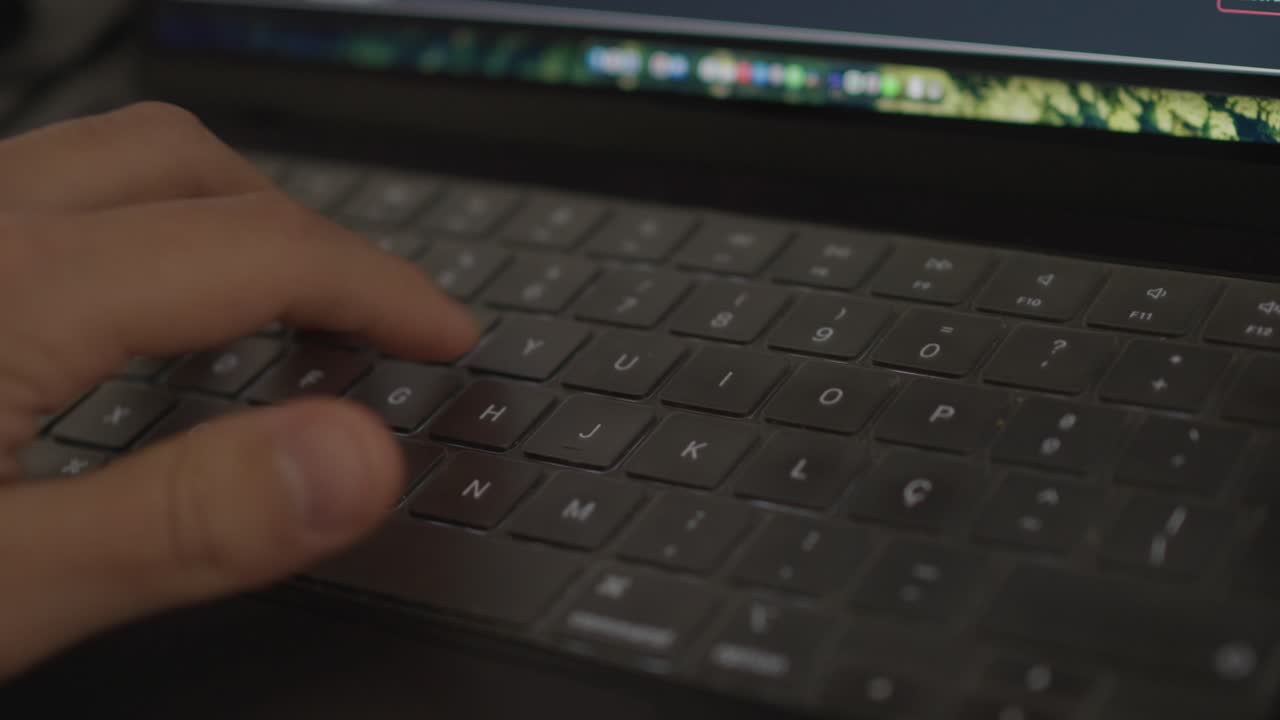 Primo piano della mano che digita sulla tastiera di un laptop con il bagliore dello schermo in un ambiente di lavoro interno scarsamente illuminato.
