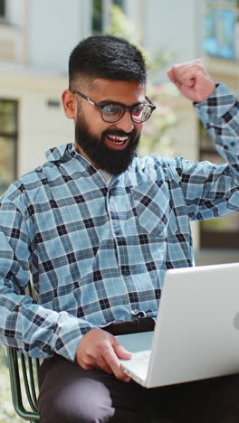 Glücklicher-Indischer-Mann,-Der-Am-Laptop-Arbeitet,-Feiert-Erfolg-Und-Gewinnt-Geld,-Während-Er-Auf-Einer-Städtischen-Straße-In-Der-Stadt-Sitzt