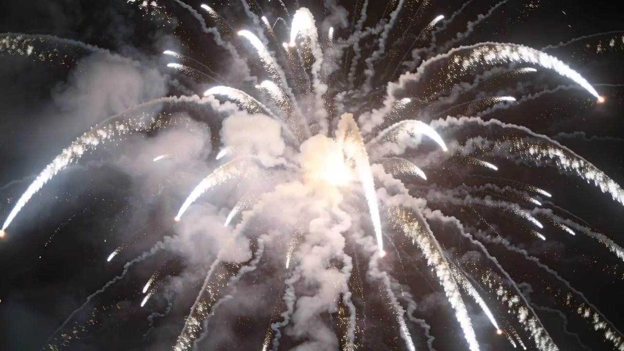 Estallido de fuegos artificiales blancos encendiéndose y expandiéndose durante un espectáculo nocturno, produciendo largas estelas de chispas y humo.