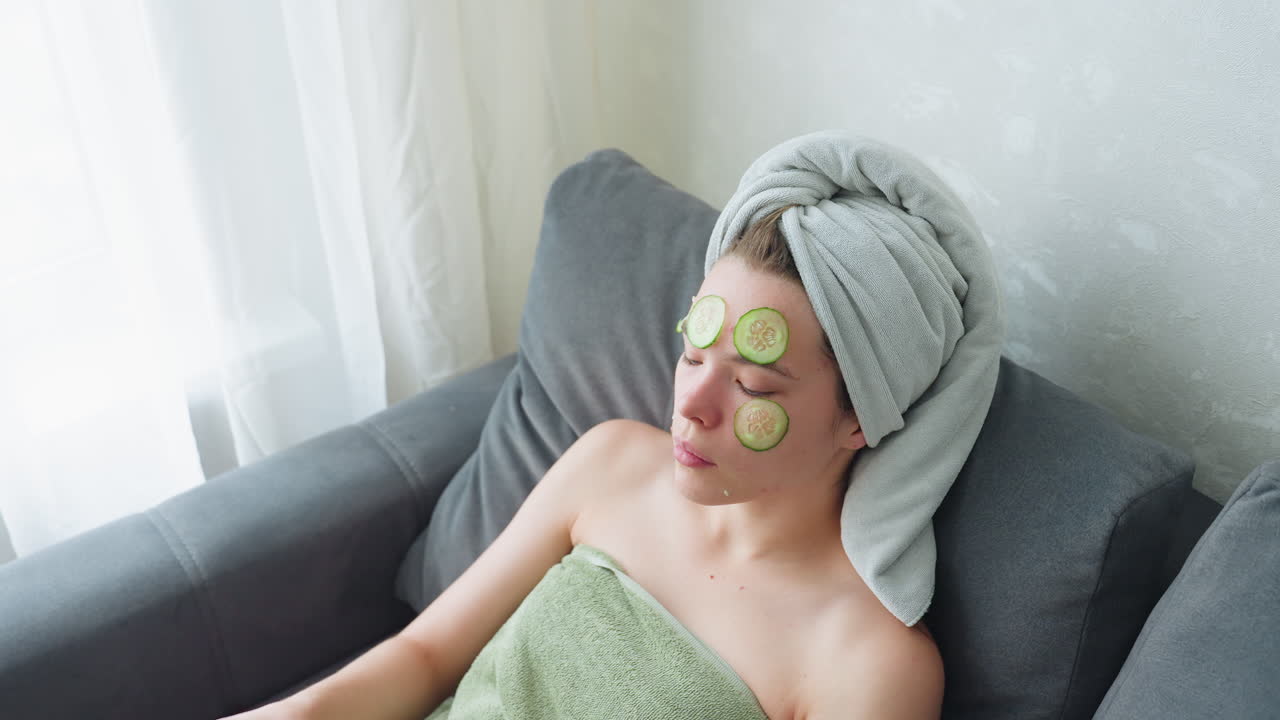 Young lady relaxing on couch beside window applying cucumber slice on cheek for refreshing beauty treatment, skincare ritual for glowing skin, soft light adding calm, rejuvenating atmosphere