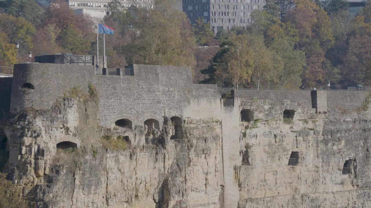 Ancient stone fortress wall with arched openings, surrounded by autumn trees and modern buildings