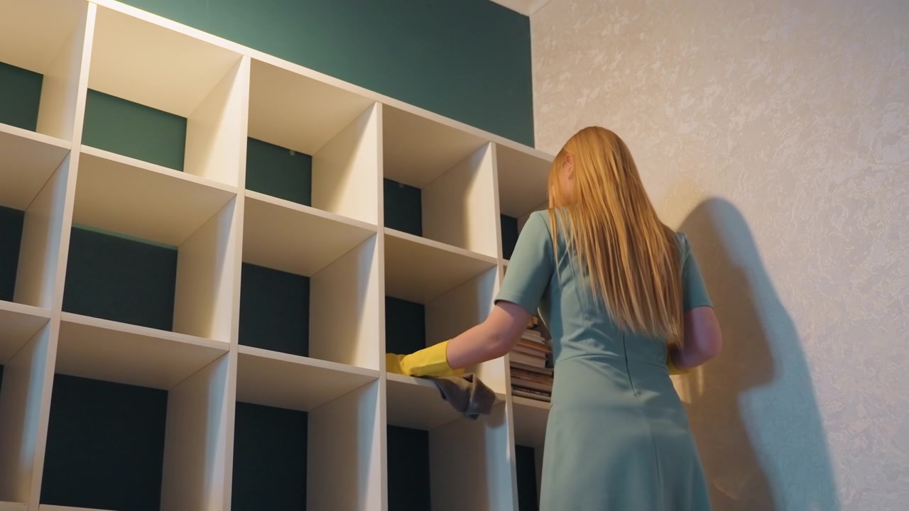 Donna che pulisce a fondo gli scaffali di casa, indossando guanti gialli e tenendo una borsa di tela, pulendo scomparti impolverati contro una parete color verde acqua, con un umore tranquillo e determinato, preparandosi per un trasloco e uno sgombero.