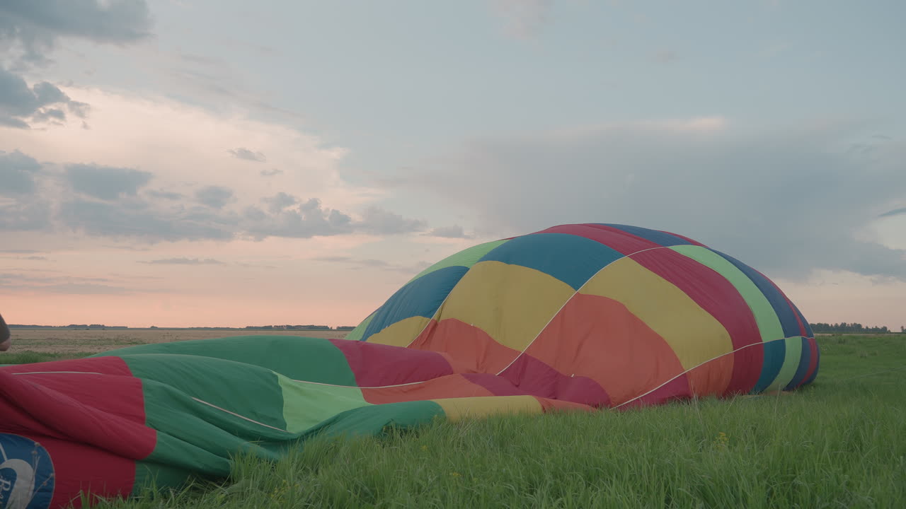 man holding rope while colorful hot air balloon envelope fills with pumped air on grassy open field during sunset preflight setup teamwork and adventure mood with vibrant sky