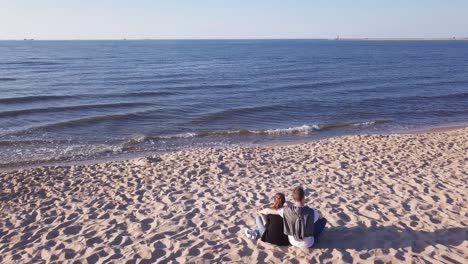 Toma-Aérea-De-Una-Pareja-Joven-Sentada-En-La-Playa-De-Arena,-Abrazándose,-Tomándose-De-La-Mano