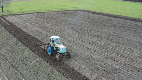 Vista-Aérea-De-Viejos-Equipos-Tractores-Cultivan-Tierras-De-Cultivo,-Emiten-Gases-De-Escape-Azules