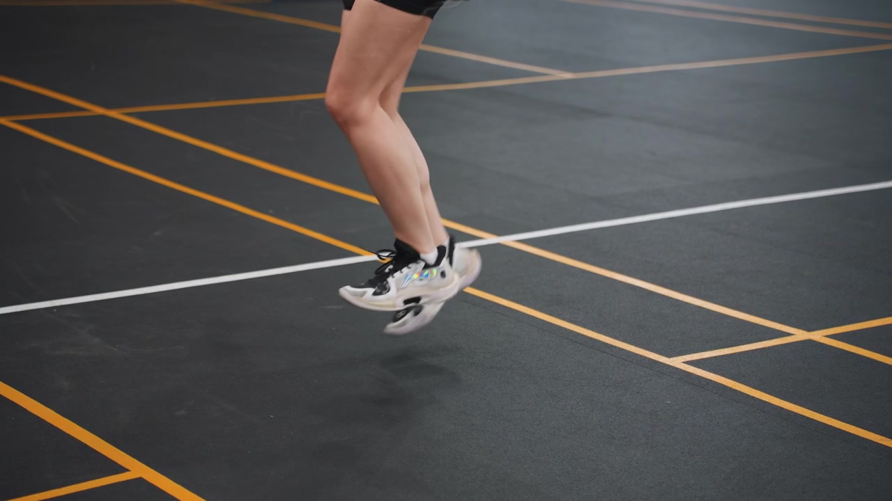 Linee del campo, giocatore bianco si allena nel badminton con spostamento laterale, posizione bassa e pivot rapidi, superficie del campo gommata con segnaletica gialla, focus su velocità e tempismo, sessione tecnica guidata dall'allenatore.