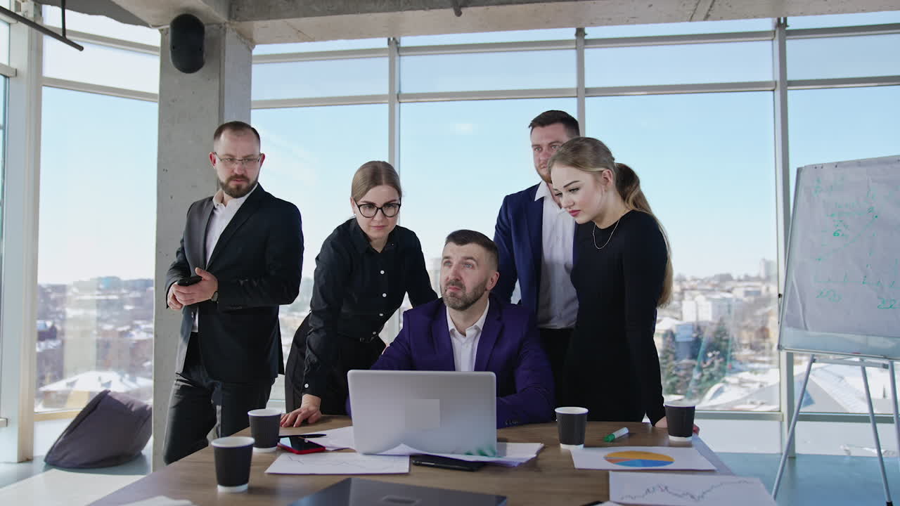Office team standing around desk with laptop. Another man joins them after phone conversation. Sunny cityscape at the backdrop.