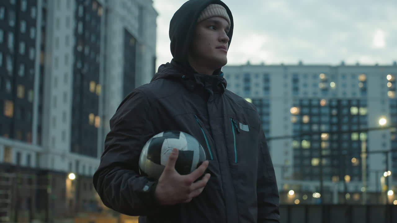 Young man holding soccer ball standing confidently in modern sport arena, surrounded by tall residential buildings under dramatic evening sky, lost in thought