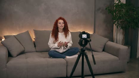 Porträt-Einer-Langhaarigen-Schönen-Frau,-Die-Auf-Einer-Grauen-Großen-Couch-Mit-Hintergrundbeleuchtung-Im-Wohnzimmer-Im-Loft-Interieur-Sitzt-Und-In-Die-Kamera-Spricht.-Junge-Bloggerin-Macht-Ein-Video