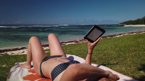 Eine-Aufnahme-Einer-Frau,-Die-Ein-Ebook-An-Einem-Grünen-Strand-Auf-Einer-Insel-Liest