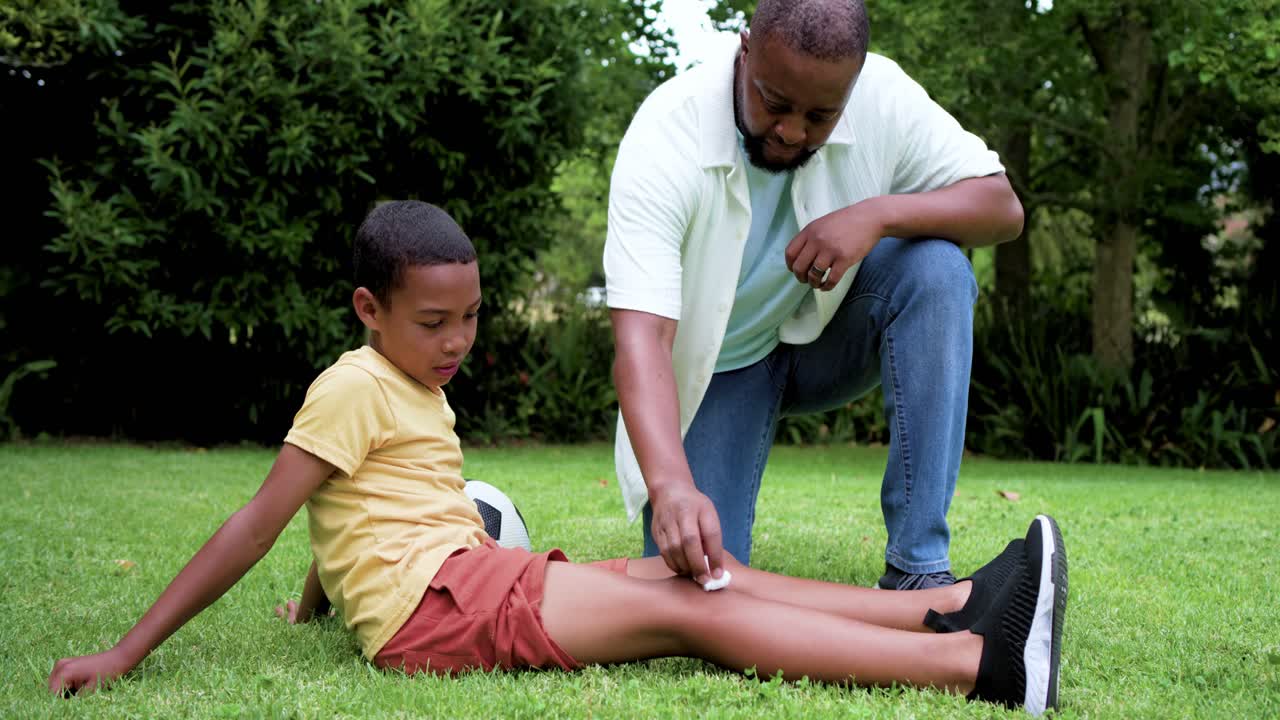 African American father kneeling, dabbing and bandaging school-age son's shin after scrape on lawn