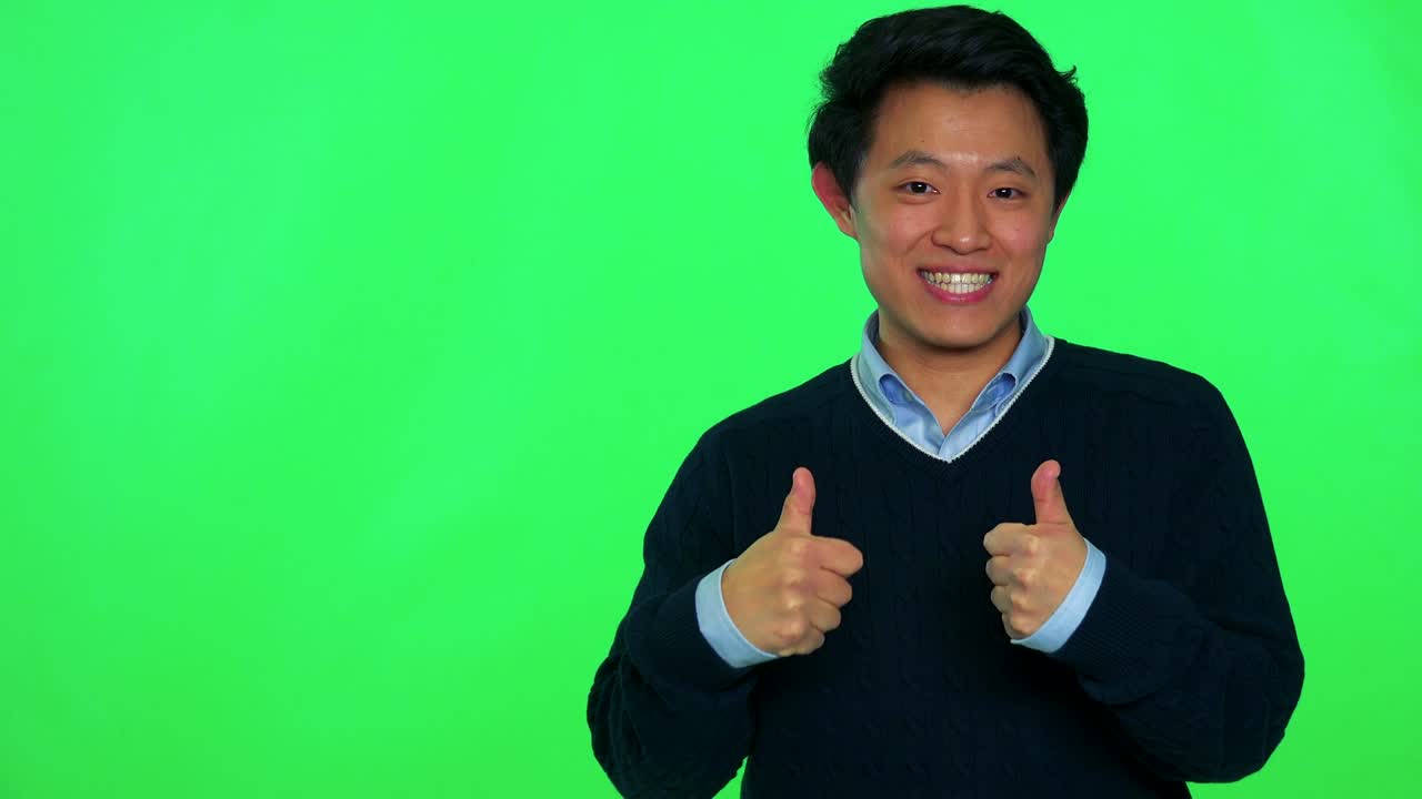 un joven asiático sonríe y muestra los pulgares hacia la cámara - estudio de pantalla verde