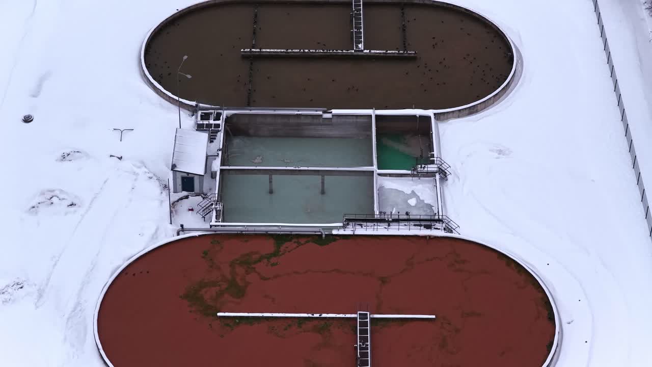 Top-down drone shot of snow-covered wastewater treatment clarifier basins at an industrial facility, showing settling tanks and process ponds in harsh winter conditions