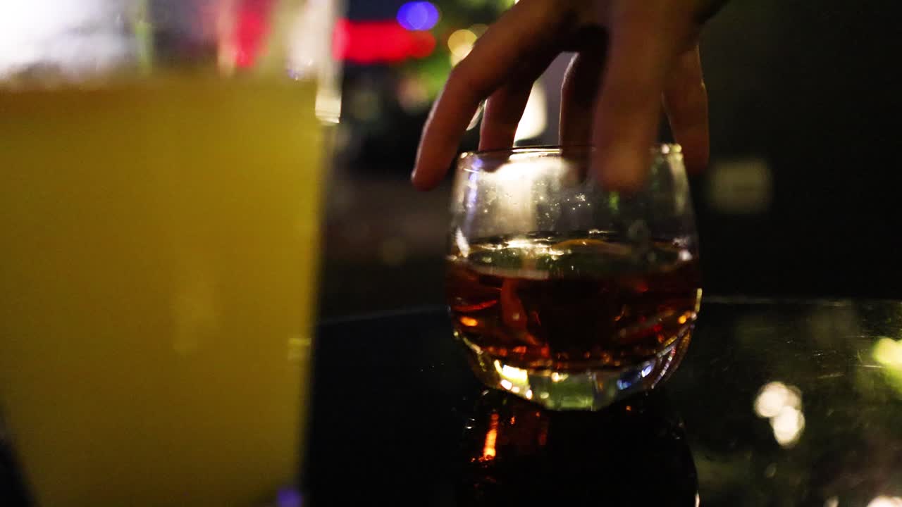 A hand reaches for and lifts a cocktail glass from a reflective bar counter, with dim ambient lighting and colorful bokeh in a nightlife setting