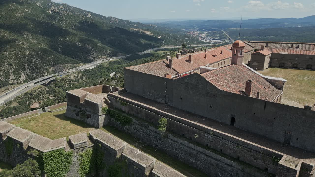 Stone walls and red tile roofs make up fort bellegarde, a historic fortress in the french countryside. Lush green valleys and a winding road can be seen in the distance
