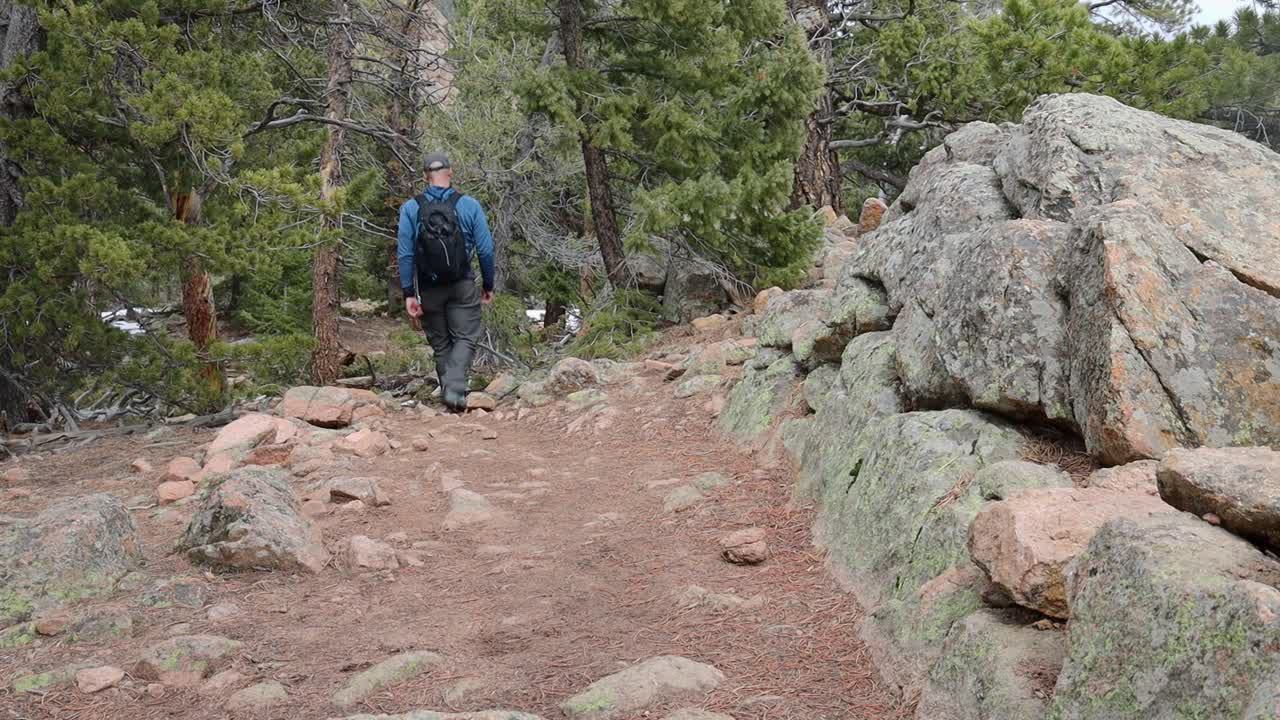 One male hiker with a backpack walking down a steep trail and by a rock wall with snow on the ground. Filmed in Staunton State Park during the spring.