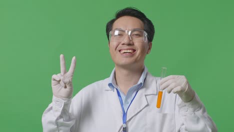 close up of asian man scientist with orange liquid in the test tube smiling and showing peace gesture while standing on the green screen background in the laboratory