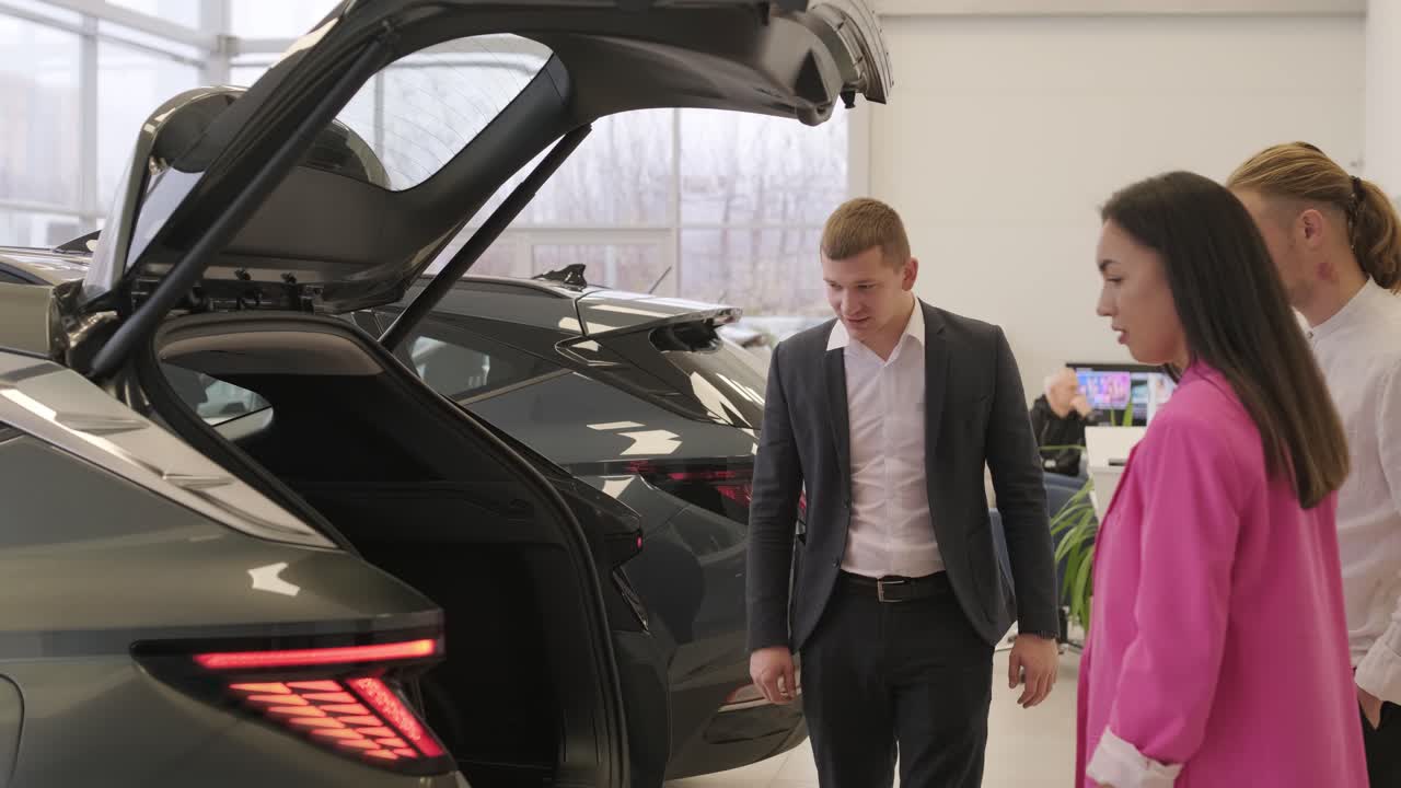 un joven vendedor de coches mostrando a una pareja de jóvenes un nuevo automóvil en el salón de concesionarios.