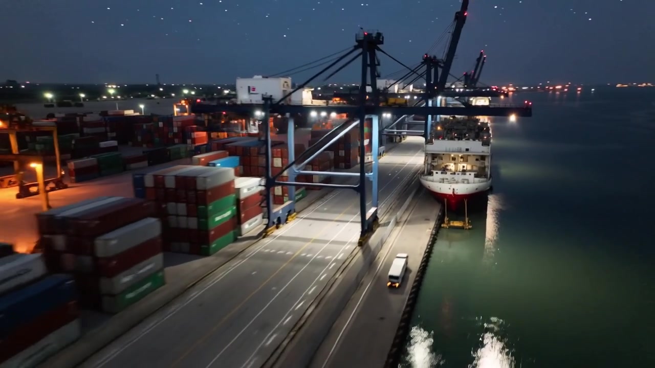 Eine sich nähernde Drohne filmt den Kai und zeigt Portalkräne, die bei Nacht ein festgemachtes Containerschiff beladen.