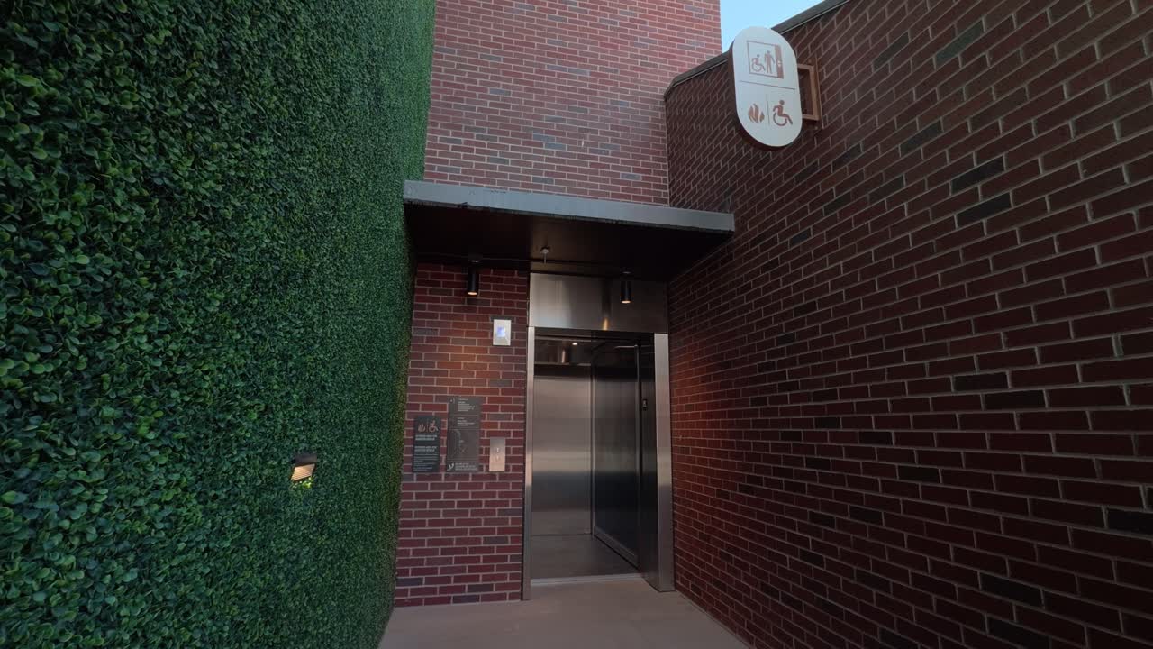 Statisk realtidsoptagelse af elevatordøre, der åbner i en overdækket murstensgang med hæk.