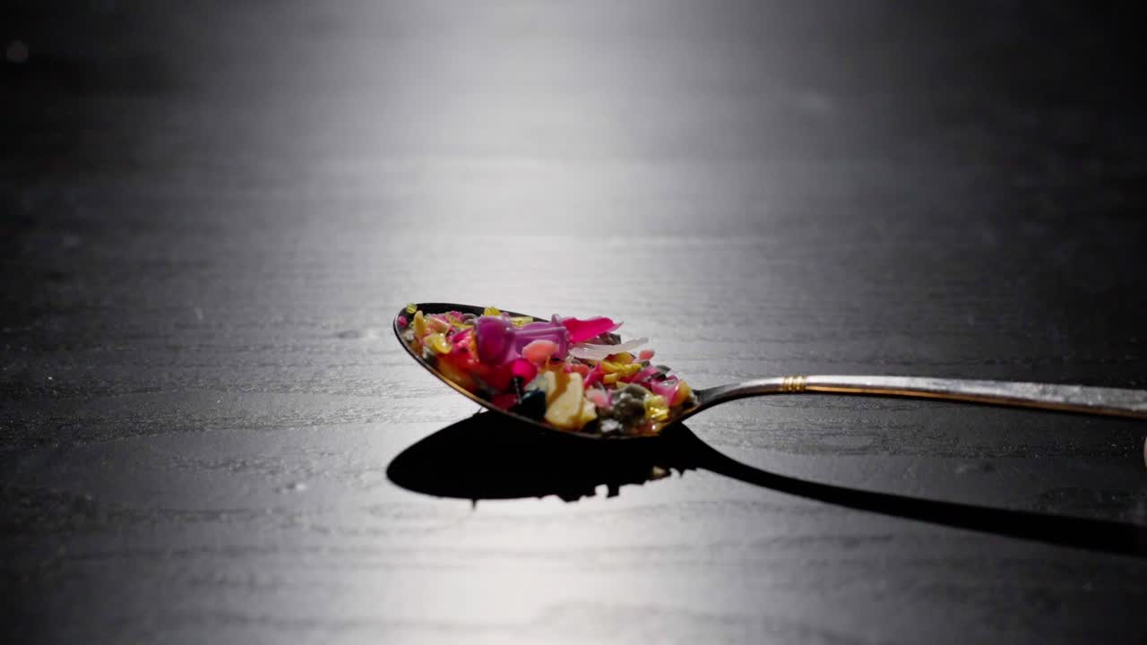 Hand tilts a spoon of colorful microplastic parts on black table. Dramatic lighting