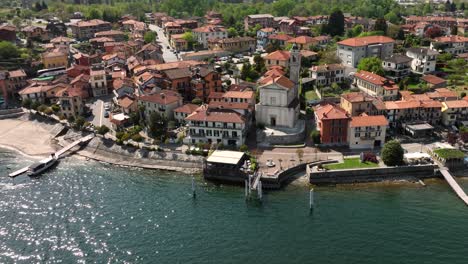 aerial slow orbit shot of an italian village on the shore of lago maggoiore on a bright sunny day with shimmering waters, and classic tile roof buildings