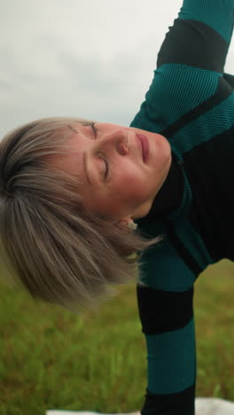 vista en primer plano de una mujer avanzada en traje verde-negro de pie en una alfombra de yoga, practicando la postura de triángulo de yoga con los brazos abiertos y las piernas separadas, puesta contra un cielo nublado en un tranquilo campo de hierba