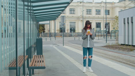 young woman with face mask using smartphone at a modern tram stop