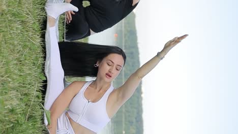 two beautiful slender young women are doing yoga she is sitting on a mat and raising her arms up two beautiful slender young women are doing yoga she is sitting on a mat and raising her hands up in the city park women practicing yoga outdoors on a sunny day