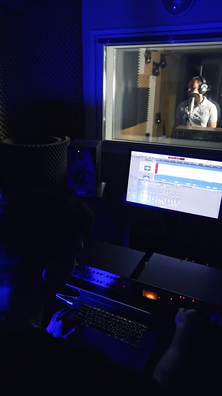 Music producer working in the little dark recording room. Sound engineer looks at computer screen. Male singer performing a song at backdrop. Vertical video