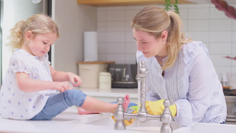 Madre-Usando-Guantes-De-Goma-En-Casa-En-La-Cocina-Con-Su-Hija-Pequeña-Divirtiéndose-Mientras-Lavan-En-El-Fregadero,-Filmada-En-Cámara-Lenta