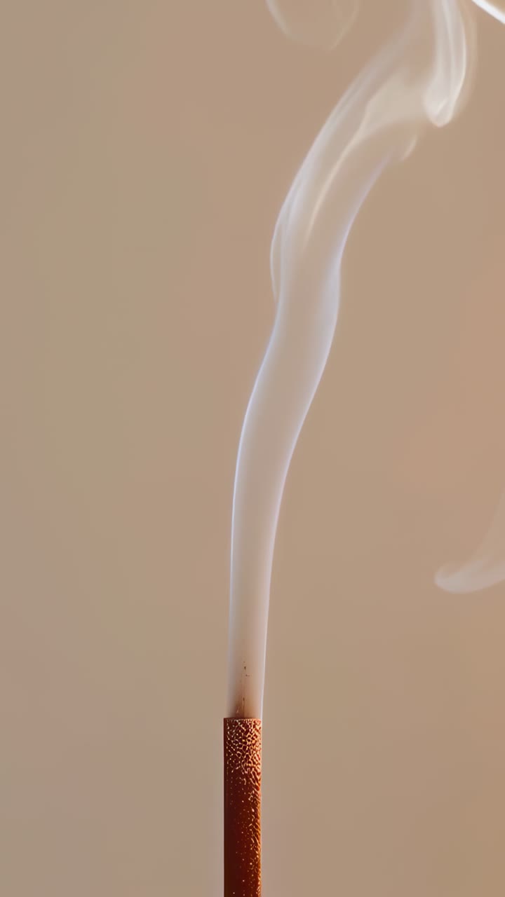 Vertical video: Showing lit incense stick emitting curling smoke over beige backdrop, glowing tip