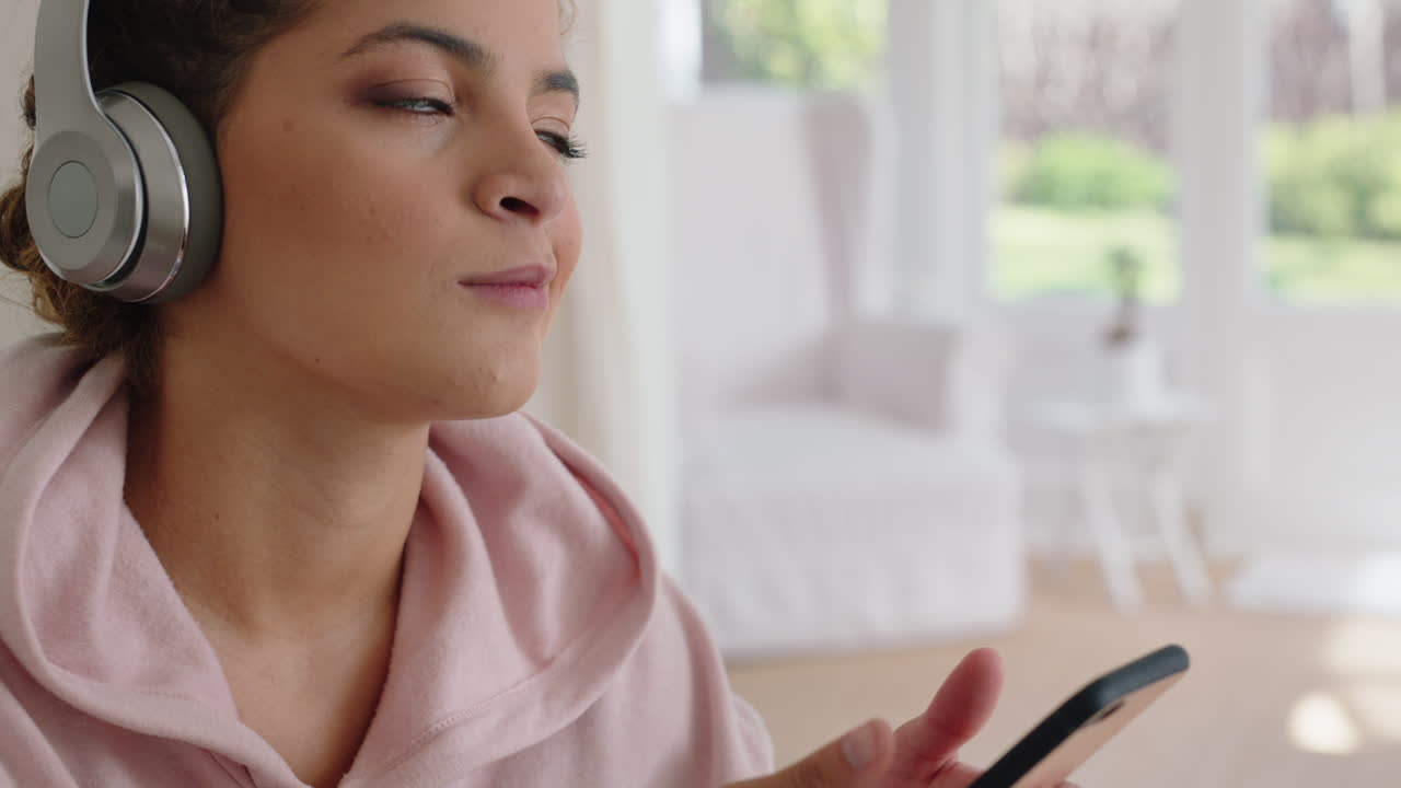 아름다운 십대 소녀가 음악을 듣고 헤드폰을 착용하고 스마트폰으로 문자를 보내고 집에서 휴식을 취하고 주말 아침에 소셜 미디어에서 라이프 스타일을 공유하는 것을 즐기고 있습니다.