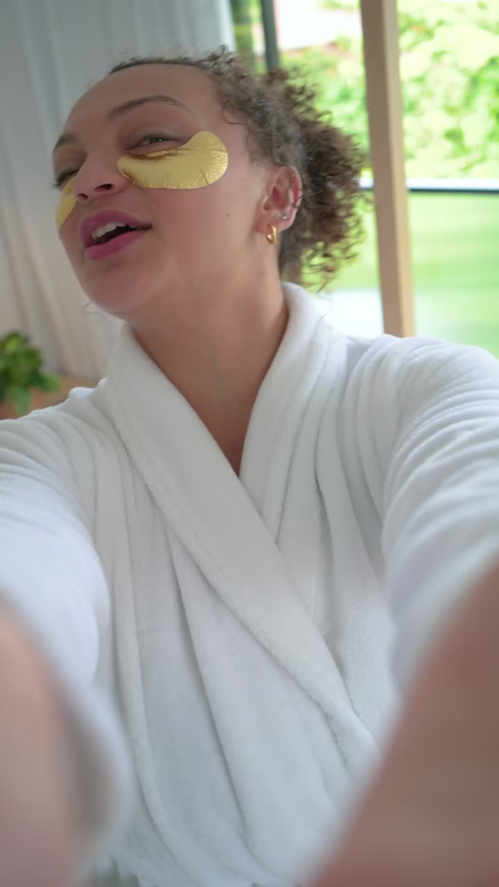 Vertical video: Woman wearing robe, starting selfie in bedroom, showing gold patches for skincare