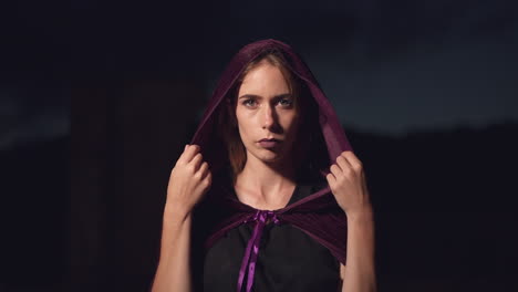 mysterious woman wearing a purple cape slowly putting the hood over her head and staring at the camera