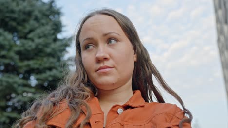 Low-angle-looking-up-at-beautiful-brunette-woman-shrugging-with-no-care,-cloudy-sky-and-tree-out-of-focus