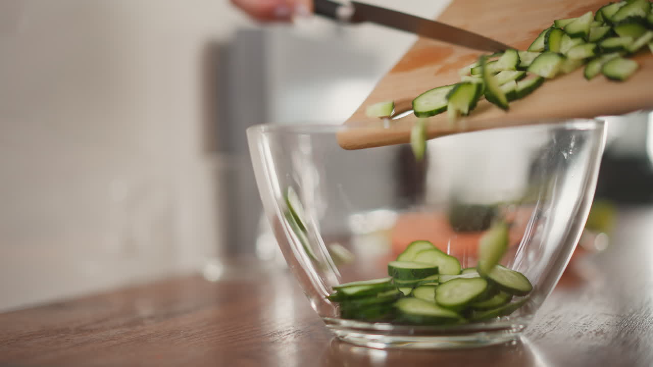 Transparent glass bowl on wooden counter as freshly sliced cucumber is poured from cutting board into bowl, capturing clean food preparation moment in soft natural kitchen lighting