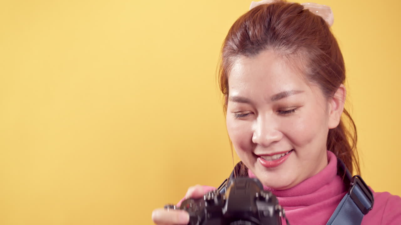 Young Asian woman playing in pink clothing using a digital camera against an isolated yellow background
