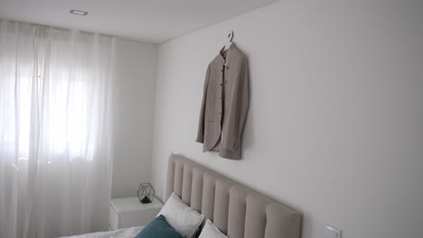 beige wedding suit hanging on white bedroom wall, soft light from window, modern minimalist decor