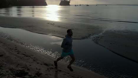 aerial tracking shot of silhouetted young man jogging on a beach in auckland, new zealand